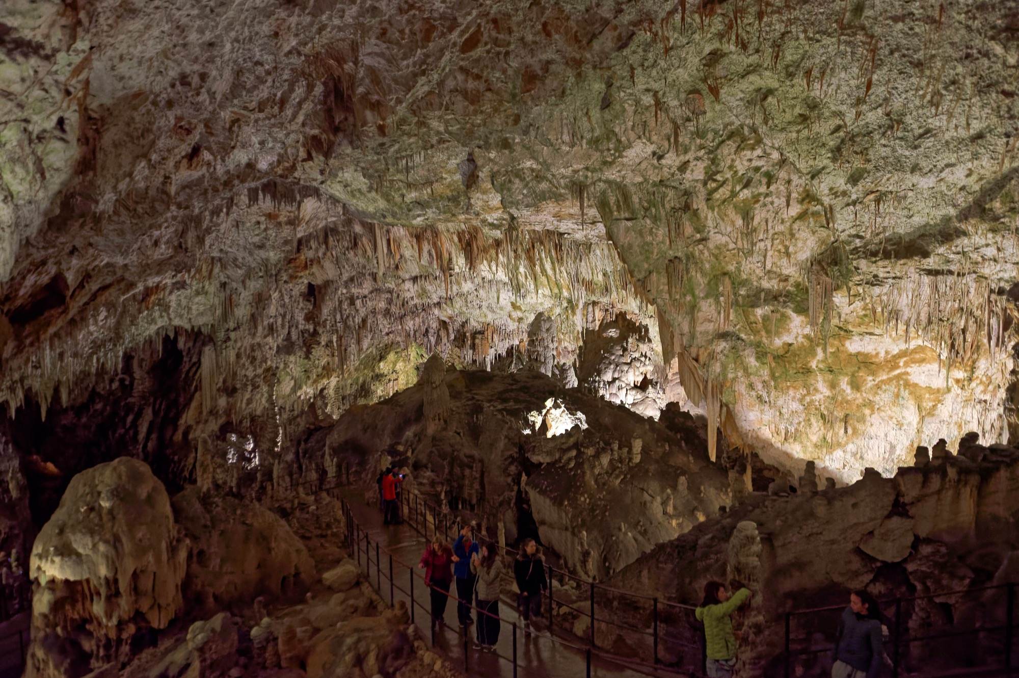 A railed footpath winding between stalagmites under a ceiling of stalactites in Postojna Cave